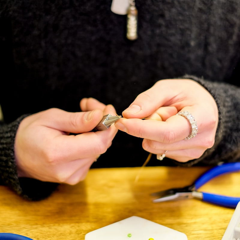 Mains travaillant sur un bijou avec des pinces et du fil.